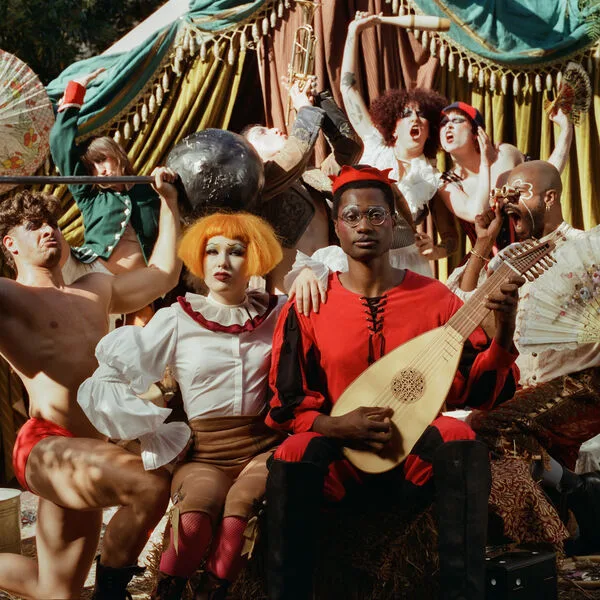 The Big Disgrace album cover features Haute and Freddy seated in front of ornate curtains, surrounded by dancers. Freddy holds a musical instrument, while the performers strike dramatic, theatrical poses. The aesthetic is distinctly carnivalesque, combining pastel tones with flashes of vivid red, heavy makeup, brightly colored hair, and exaggerated styling. The overall composition evokes a flamboyant stage performance, blending camp, theatricality, and playful excess.